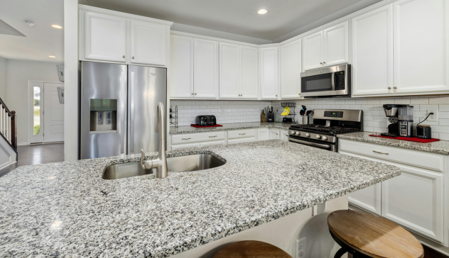 A stylish kitchen with white matte cabinets and polished granite countertops that create an elegant contrast. The glossy finish of the countertop complements the smooth texture of the cabinets, adding depth to the overall design. The open layout and neutral tones give the kitchen a bright and spacious feel, while the natural stone texture of the countertop enhances the visual appeal and brings a touch of sophistication. This combination of matte and polished finishes helps balance functionality and aesthetics in this modern kitchen.