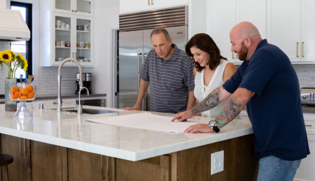 A couple works with a kitchen designer at the island, reviewing kitchen plans. The light-filled space features a spacious kitchen island and modern appliances, demonstrating the collaborative approach to designing a kitchen that fits the homeowners' needs.